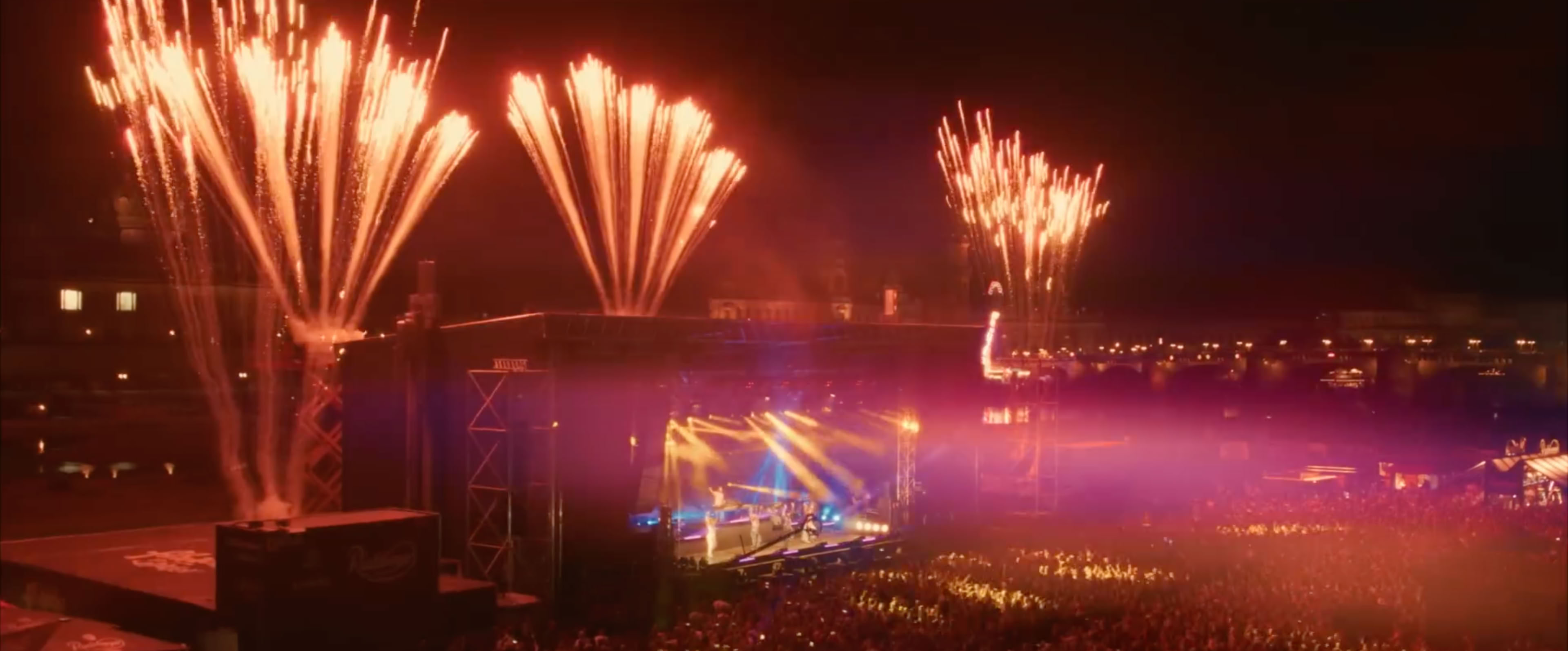 Feuerwerk über einer beleuchteten Konzertbühne mit einer großen Menschenmenge im Vordergrund.